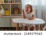 Small photo of A little girl cries and is naughty sitting at the table in the nursery. Children's tantrums, tears and discontent. Problems of kid's upbringing.