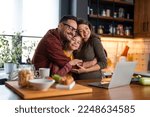 Small photo of Loving family of three embracing in the kitchen. Mom and dad are daydreaming with the eyes closed while little girl is smiling and looking at the camera. They're standing at the kitchen counter.