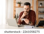 Small photo of Serious business man sitting at table in home office, holding and looking at credit card, worried about debt and credit card deficit, considering a financial loan from bank.