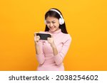 Beautiful young asian women play mobile game and put on wireless headphone standing on isolated yellow background. Playing game on smartphone winning victory moment. Very enjoy and fun relax time