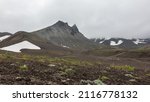 Small photo of A picturesque mountain, devoid of vegetation, against a cloudy sky. Patches of melted snow on the slopes. Scant plants grow in the valley. The path winds through the hills. Kamchatka. Mount Camel