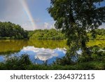 a stunning shot of the silky brown lake water at Candler Lake with green trees and plants reflecting off the water with blue sky, clouds and a rainbow at Lullwater Preserve in Decatur Georgia
