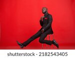 Creative emotional Portraits of a young mid-adult Kenyan black Male Man wearing a black complete suit black tie and red pocket size, black socks, and shoes against a red background in the studio