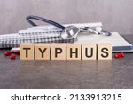 Small photo of TYPHUS - text is made up of wooden blocks standing on a gray table. In the background is a stack of paper notebooks for writing and tablets. medical concept, gray background