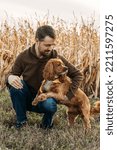 Small photo of Autumn portrait of a handsome caucasian man wearing a brown knit sweater and jeans. He is squatting down beside his cocker spaniel dog. There is a corn field behind them. This was taken in Canada.