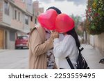 Two people nearby holding heart-shaped balloons in the street and surrounded by some plants. Meaning love, affection, lovers, among others.