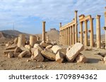 The ruins of an ancient temple, consisting of columns in perspective and marble stones in the foreground, in the city of Palmyra, under a blue sky and a mountain with a fortress on top, on the horizo