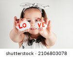 Small photo of Smiling little girl in white clothes hold two white smooth stones with syllables lo and ve painted red on white background. Family, love, birthday, Valentine's day, Mother's day concept. Copy space.