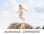 Small photo of Happy little girl jump up at top of haystack with hands to sides and look down. Joyful kid having fun on hayrick. Outdoor walking. Beautiful blue sky. Freedom. Summer vacation, countryside lifestyle.