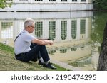 Small photo of Elderly gray-haired man sits on the shore of a city pond and holds a smartphone in his hands. Recreation of townspeople on a summer day. An elderly man uses a smartphone. Moscow, Russia - 08.13.2022