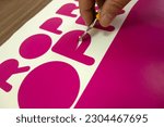 Small photo of The hand of a man with a stationery knife, selects a vinyl film after cutting on a plotter.Sampling of the film in the production of avtovinil. Cutting a pink sticker.