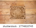 Opened gift box with shredded paper on wooden background. Brown paper box with decorative straws fillers for your product placement. Flat lay, top view. Centre composition with empty space for produce