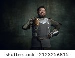 Small photo of Tasting beer. Happy medieval warrior or knight with dirty wounded face holding big mug of beer isolated over dark vintage background. Comparison of eras, history, renaissance style, festival, ads