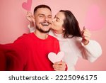 Portrait of lovely young couple, woman kissing happy and smiling man, taking selfie together isolated over pink background. Concept of love, relationship, Valentine's Day, emotions, lifestyle