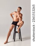 Small photo of Full-length portrait of handsome, muscular young man sitting on chair in underwear and posing against grey studio background. Concept of men's health and beauty, body care, fitness, wellness, ad