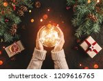 Female hands holding Christmas glowing ball on holiday background with Fir branches, gifts. Xmas and Happy New Year card, bokeh, sparking. Flat lay, top view