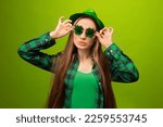 Small photo of Haughty stylish young woman wearing party green hat, clover shaped glasses, checkered plaid shirt and looking aside isolated on green background. St Patricks Day or March 17 celebration concept.
