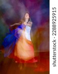 Small photo of Mystical, blurred image of a female professional dancer in a white dress, dancing with floating, red and blue, gauzy fabrics in a studio, shot against a gray background.
