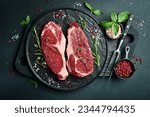 Two raw rib eye beef steaks with spices on a plate. Ready to cook. On a dark stone background. Top view.