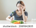 Small photo of Diet in bored, unhappy beautiful asian young woman, girl on dieting, holding fork at broccoli in salad plate, dislike or tired with eat fresh vegetables. Nutrition of clean, healthy food good taste.