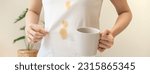 Small photo of Cloth stain, disappointment asian young woman clumsy with hot coffee, tea stains on shirt, hand show making spill drop on white t-shirt, spot dirty or smudge on clothes at home, isolated on background