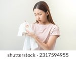 Small photo of Housewife, asian young maid woman hand in holding white T-shirt, looking at cloth stain, spot dirty or smudge on clothes, dirt stains for cleaning before washing, making household working at home.