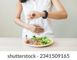 Intermittent fasting, health care asian young woman dietary, having stomach ache, temptation hungry of breakfast food in morning on table at home, looking at watch on her wrist but not yet time to eat