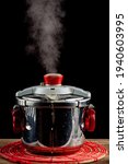 Small photo of A steel pressure cooker is cooling on a fabric trivet on wooden table. The pressure valve is released and steam is coming out as the system gets depressurized. Hot steam leaving pot is seen.