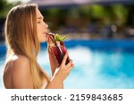 Pretty woman drinks refreshing cocktail decorated with mint, cherry and lemon. Female enjoys a drink near swimming pool with blue water. Girl chilling with beverage in tropical sun. Vacation concept