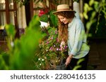 Small photo of Young woman standing in garden planting and watering flowers in pots smiles. Junior female gardener with gardening tool in orchard taking care of potted plants. Farming and gardening concept