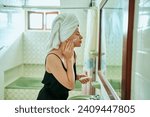 Woman in a vintage bathroom applies sunscreen to her face for skin protection. Towel-wrapped hair, retro tiles, skincare routine, mirror reflection capture the essence of daily beauty regimen.
