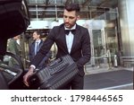 Small photo of Considerate hotel porter wearing uniform handling a piece of luggage and placing it into a car trunk