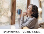 A beautiful girl in a sweater drinks tea or coffee against the background of a window and lights. Christmas or fall time