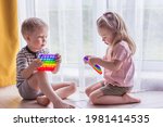 Blonde boy and girl children sit on the floor near the window and play silicone toy antistress pop it. Pop it sensory toy. Stress relief. Colorful anti stress silicone sensors toy