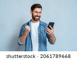 Small photo of Happy satisfied man looking at mobile phone screen gesturing yes with clenched fist isolated on blue studio copy space. Overjoyed excited joyful guy make winner gesture read message about reward