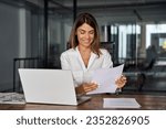 Small photo of Smiling European Latin business woman accountant analyst holding documents, work at laptop computer doing online trade market tech research. Focused Hispanic businesswoman with paperwork in office.