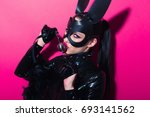 Beautiful dominant brunette vamp mistress girl with fashion makeup in glamour latex dress, collar and bdsm black leather fetish rabbit mask posing on hot pink backgroung