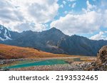 Lovely autumn landscape with turquoise mountain lake against high mountain top in sunlight under clouds in blue sky. Sunlit beautiful small alpine lake and large mountain range. Vivid autumn colors.