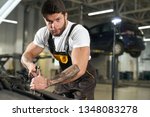 Small photo of Muscular mechanic in uniform repairing car in autoservise station. Handsome, brutal bearded repairman with dark hair and tattoos on hands holding wench and looking at camera.