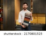 Small photo of Confident male expert presenting variety samples of upholstery fabric in dark lightened showroom. Portrait of smiling brunet salesman showing colorful textile palette. Concept of interior design.