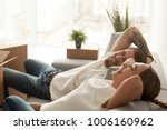 Happy couple relaxing on sofa having fun on moving day, excited young homeowners enjoying relocation in into new home, positive laughing man and woman sitting on couch with packed cardboard boxes