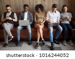 Multicultural young people using laptops and smartphones sitting in row, diverse african and caucasian millennials entertaining online obsessed with modern devices waiting in queue, gadget addiction