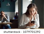 Small photo of Young curious guy likes millennial beautiful girl looking with interest watching smiling lady sitting nearby in cafe using smartphone, flirt in public place, dating and love at first sight concept