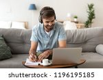Man sitting on couch in living room at home enjoying studying using laptop and headset looking at device screen listening audio making some notes. Male has lesson online e-learning in internet concept