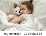 Small photo of Cute little kid girl hugging teddy bear sleeping lay in cozy bed, happy small child embracing toy fall asleep on soft pillow white sheets covered with blanket having healthy night sleep, top view