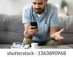 Close up confused man having problem with broken not working phone, annoyed frustrated male student receiving bad news, reading unpleasant email, sitting on sofa, looking at screen, spam message