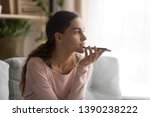 Woman sitting on couch holds phone talks on speakerphone with friend, makes voice recognition or request uses internet services through virtual assistant, record audio message, translator app concept
