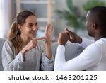 Smiling mixed ethnicity couple or interracial friends talking with sign finger hand language, happy two deaf and mute hearing impaired people communicating at home sit on sofa showing hand gestures