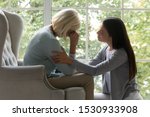 Empathic millennial lady soothing desperate crying mature mother, sitting in armchair. Side view upset grown up daughter comforting, supporting caring unhappy middle aged mommy felling unwell at home.