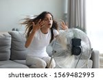 Small photo of In living room without air-conditioner tired from summer heat young woman turned on floor ventilator waving her hands to cool herself, female sitting on couch suffers from unbearable too hot weather